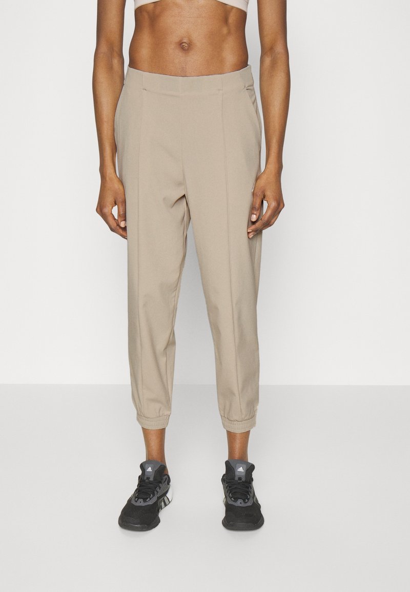 Beige, zulaufende Hose mit elastischen Bündchen, hergestellt aus einem leichten Stoff. Verfügt über einen Bund mit vorderen Falten. Kombiniert mit schwarzen Sportschuhen.