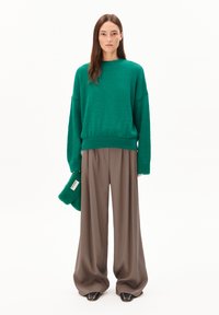 Frau trägt einen übergroßen grünen Pullover, weite braune Hosen, schwarze Nieten-Schuhe und hält einen passenden grünen Strickhut vor einem weißen Hintergrund.