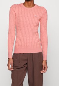 Pull rose clair à maille torsadée avec un col et des poignets côtelés, arborant un petit emblème sur la poitrine. Texture douce, coupe ajustée.