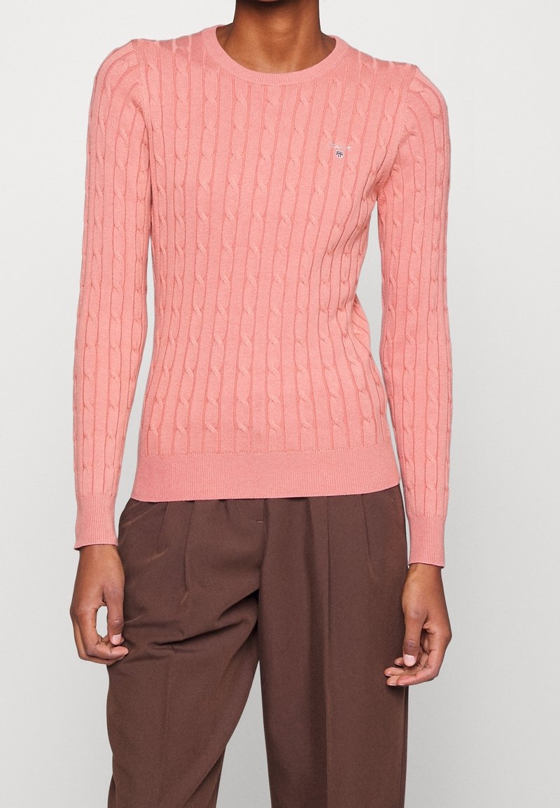 Pull rose clair à maille torsadée avec un col et des poignets côtelés, arborant un petit emblème sur la poitrine. Texture douce, coupe ajustée.