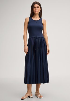 Frau steht und trägt ein ärmelloses, marineblaues, plissiertes Midi-Kleid und weiße flache Sandalen mit Riemchen vor einem einfarbigen hellen Hintergrund.