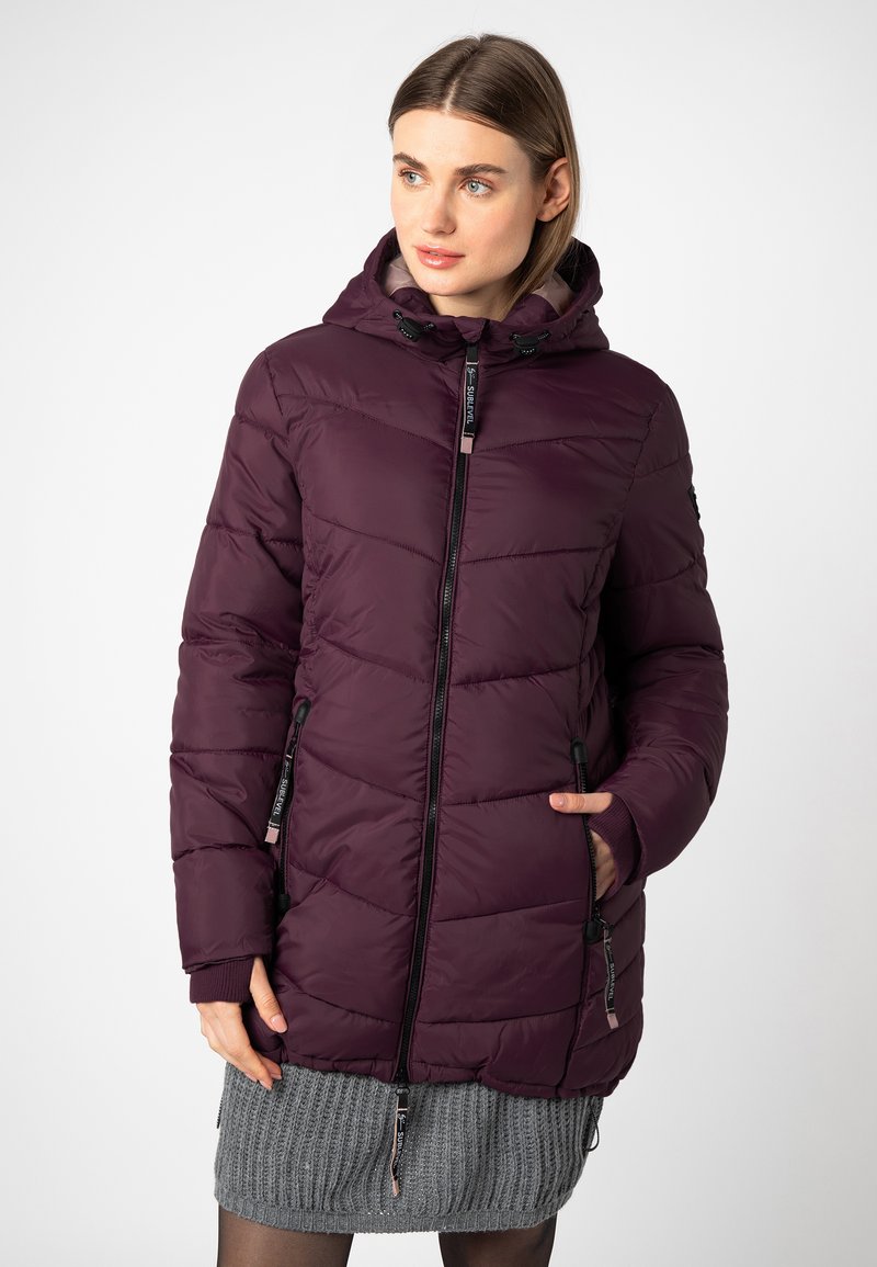 Vrouw in een donkerpaarse winterjas met capuchon en gewatteerd, met ritszakken, en een grijze gebreide rok tegen een witte achtergrond.
