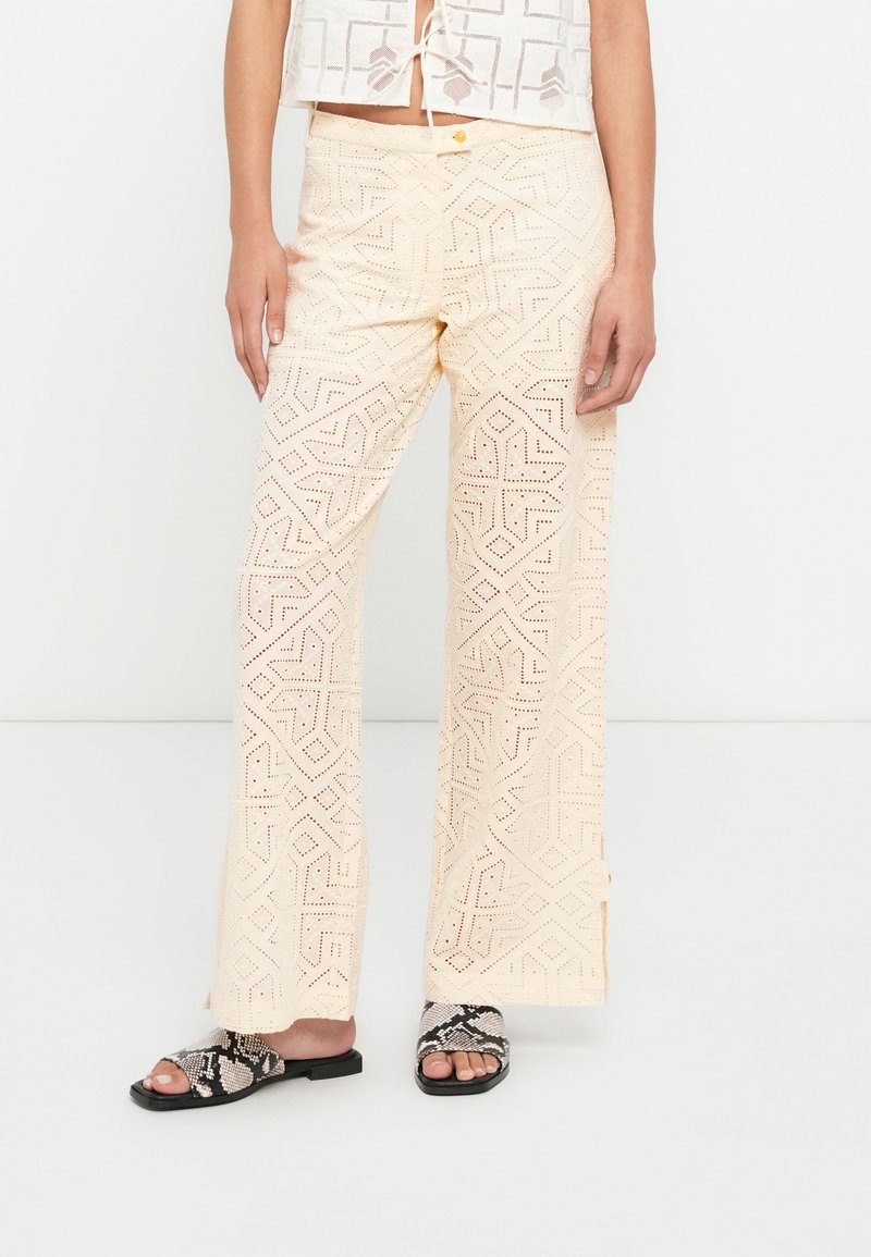 Pantalon couleur crème à jambes larges avec un motif géométrique ajouré, une fermeture par bouton et une finition texturée. Porté avec des sandales plates noires.