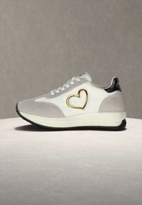 Ανδρικά αθλητικά παπούτσια σε λευκό και γκρι σουέντ με σχέδιο χρυσής καρδιάς, μαύρες λεπτομέρειες στη φτέρνα και υφασμένο καουτσούκ στην εξωτερική σόλα.