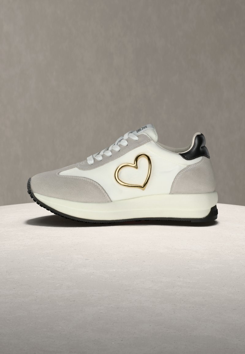 Ανδρικά αθλητικά παπούτσια σε λευκό και γκρι σουέντ με σχέδιο χρυσής καρδιάς, μαύρες λεπτομέρειες στη φτέρνα και υφασμένο καουτσούκ στην εξωτερική σόλα.