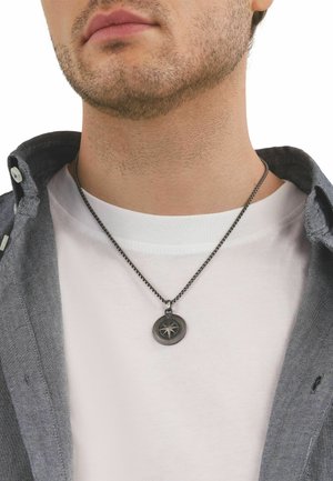 Primer plano del cuello de un hombre que lleva un colgante de brújula plateado en una cadena, camiseta blanca de cuello redondo y cuello de camisa gris desabrochado.