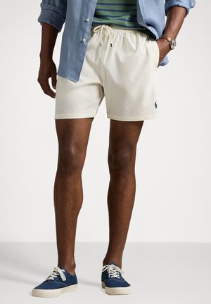 Cream-colored shorts with an elastic waistband and drawstring. Features a small embroidered logo on the left leg. Worn with a blue shirt and shoes.