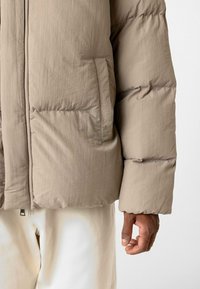 Beige pufferjack met een gladde textuur, voorzien van een zijzak, licht oversized mouwen en een rits aan de onderkant.