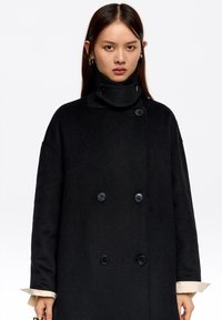 Una joven con cabello largo y oscuro lleva un abrigo negro de doble botonadura con cuello alto, con puños beige visibles en las muñecas.