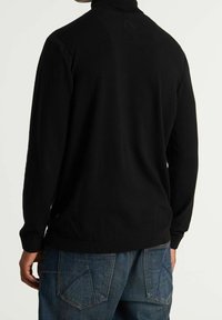 Schwarzer Pullover mit hohem Kragen, strukturierter Strickart und gerippten Bündchen. Kombiniert mit blauen Denim-Jeans mit eckigen Gesäßtaschen.