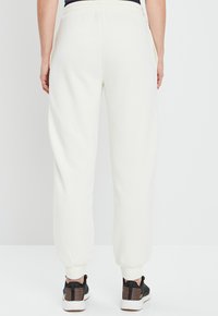 Weiße Jogginghose mit elastischem Bund und elastischen Abschlussbündchen, die eine glatte Textur aufweisen. Schwarze Turnschuhe mit gestreiften Akzenten.