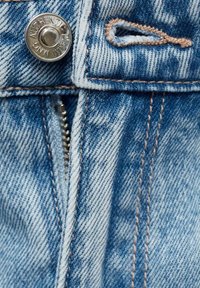 Ljusblå denimtyg med en slitagen textur, prydd med en silverfärgad knapptryckknapp, en dragkedja och kontrasterande beige sömnadsdetaljer.