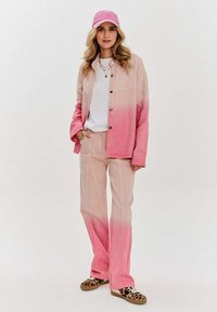 Růžový ombre kabát a kalhoty v uvolněném střihu, doplněné bílým topem a růžovou kšiltovkou; vícebarevné boty dotvářejí outfit.