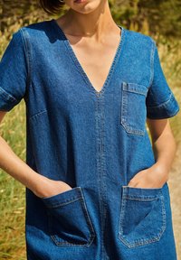 Denim haljina s dubokim V-izrezom, kratkim rukavima, dva velika prednja džepa i vidljivim detaljima šivanja, u svijetloplavoj do srednje plavoj boji.
