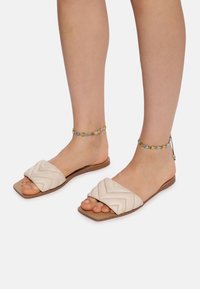 Flade slidssandaler har en lys beige læderoverdel med quiltet chevron-mønster. Sålen er brun og flad, hvilket afslører tæerne og ankelarmbåndet.
