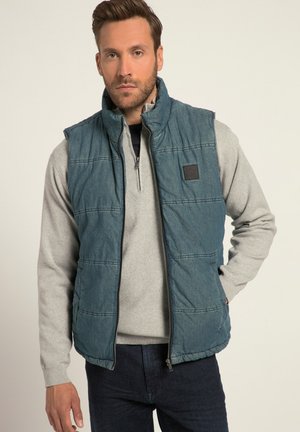 Gilet in denim imbottito di blu scuro con colletto alto, chiusura frontale con zip e due tasche laterali. Indossato sopra un maglione grigio chiaro.