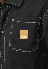 Black button-up shirt with white stitching and a chest pocket featuring a brown branded patch. The fabric appears smooth and sturdy.