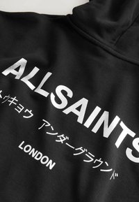 Crna kapuljača s srebrnim natpisima "ALLSAINTS" i "LONDON." Glatka tekstura i podesiva kapuljača, s modernim dizajnom ulične mode.