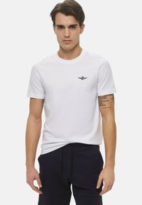 T-shirt bianca a girocollo realizzata in cotone, con un piccolo logo blu navy sul lato sinistro del petto. Maniche corte e vestibilità classica.