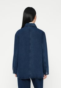 Denim jakna u tamnoplavoj boji s ovratnikom, spuštenim ramenima i dugmadi na manžetama. Glatka tekstura i ravni kroj, bez vidljivih uzoraka.