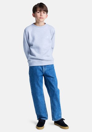 En ung gutt står med hendene bak ryggen, iført en lys grå hettegenser, blå jeans og svarte joggesko mot en enkel hvit bakgrunn.