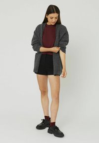 Cardigan tricoté gris foncé avec des manches retroussées, superposé à un haut bordeaux, porté avec un short noir et des chaussures noires imposantes.