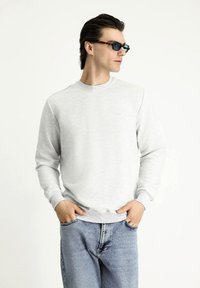 Sudadera gris claro con puños y cuello de canalé, combinada con jeans de mezclilla azul; el modelo está de pie con las manos en los bolsillos, usando gafas de sol.