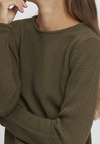 Pull en tricot texturé vert olive avec un col rond et des manches longues, présentant un motif de gaufre détaillé visible sur le tissu.