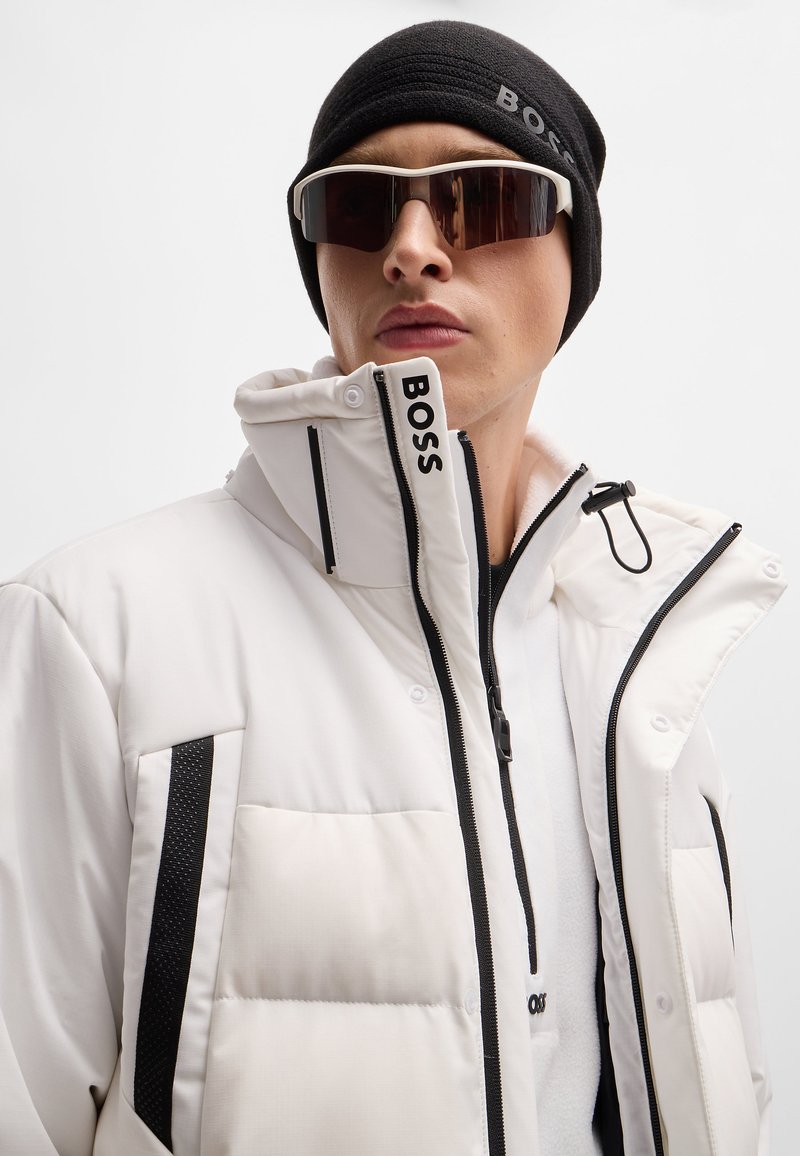 Weiße isolierte Jacke mit schwarzen Akzenten und Reißverschlüssen; hoher Kragen und Logodetails. Eine schwarze gestrickte Mütze und dunkle Sonnenbrillen vervollständigen den Look.
