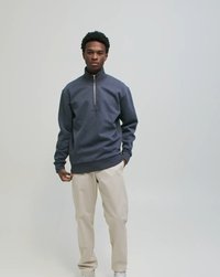 Mladi moški v mornarsko modri majici s polovičnim zadrgo, bež hlačah in belih supergah, stoji ob preprostem svetlo sivem ozadju.