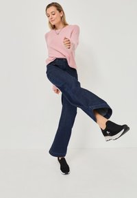 Ljust rosa, fluffig tröja med en avslappnad passform, kombinerad med mörkblå utsvängda jeans och svarta slip-on skor med vita sulor och accenter.