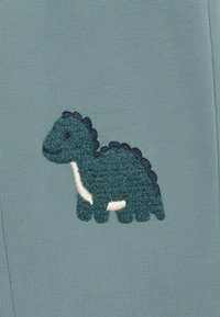 Dinosauro ricamato verde su un tessuto azzurro chiaro. Il dinosauro ha un aspetto texturizzato, con una spina dorsale blu scuro e un ventre bianco.