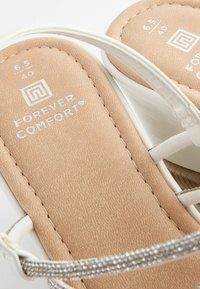Sandales blanches avec une semelle intérieure beige texturée, dotées d'une lanière argentée et de la marque "FOREVER COMFORT" embossée sur la semelle. Taille 6.5/40.