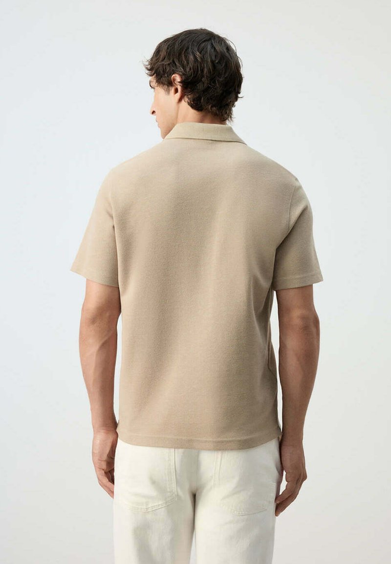 Hombre con cabello corto y oscuro, vistiendo una camisa de manga corta beige y pantalones blancos, de pie de espaldas sobre un fondo liso.