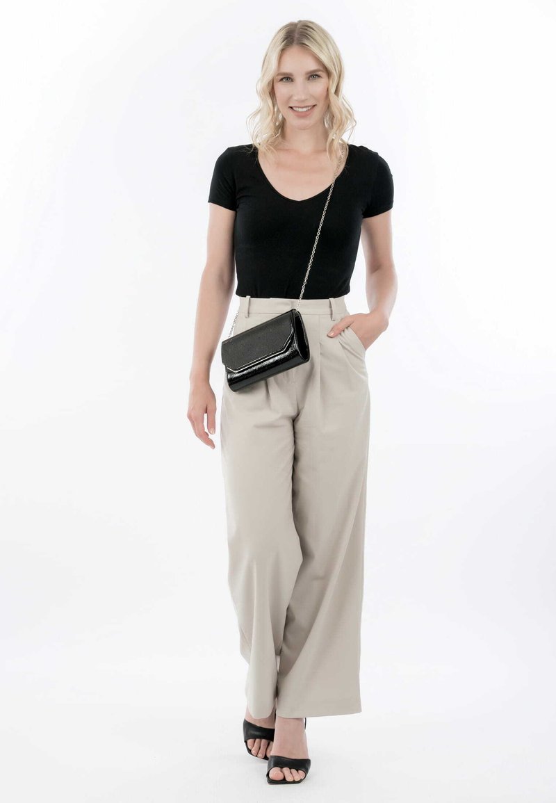 T-shirt noir ajusté, pantalon beige à jambes larges et talons ouverts noirs. Le mannequin tient un petit sac bandoulière noir avec une chaîne.