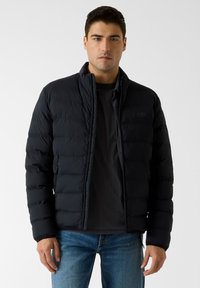 Schwarze Daunenjacke mit Stehkragen, gesteppter Optik und Reißverschluss vorne. Wird über einem schwarzen Shirt getragen und mit blauer Jeans kombiniert.