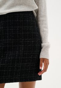 Čierna tweed mini sukňa s textúrovaným vzorom, skombinovaná s svetlošedým svetrom. Zamerajte sa na tkané detaily látky a jej tvar.