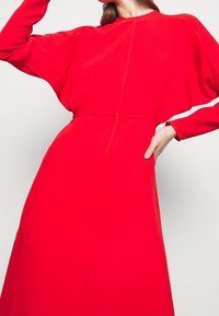 Vestido rojo de manga larga con una cintura ajustada, mangas drapeadas, textura suave y silueta recta. Detalles de costura visibles.