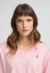 Lichtroze gebreide trui met een V-hals. Heeft een klein marineblauw logo op de linkerkant van de borst en een zachte textuur, geschikt voor casual wear.