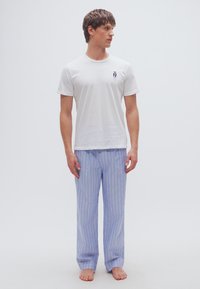 Λευκό κοντομάνικο πουκάμισο με ένα μικρό μπλε γραφικό και μπλε-λευκές ριγέ πυτζάμες. Μπαλαρίνες με γυμνά πόδια σε απλό φωτεινό φόντο.