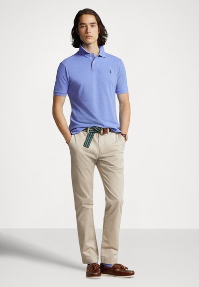 Jasnoniebieska koszulka polo z kołnierzykiem, w zestawieniu z beżowymi spodniami chinos i brązowymi butami. Dopełnieniem są wielokolorowy pasek w paski oraz zegarek.