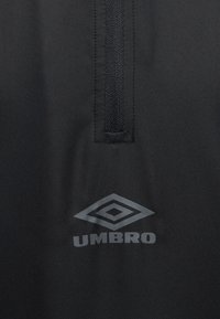Μαύρο υφασμάτινο υλικό με κάθετο φερμουάρ και ανακλαστικό λογότυπο Umbro κάτω από το φερμουάρ.