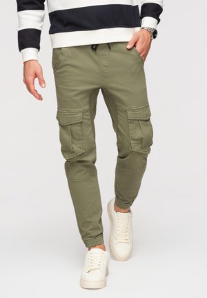 Homme portant un pantalon cargo vert olive, des baskets blanches et un sweat-shirt blanc à rayures noires, marchant vers l'avant avec une main dans la poche.