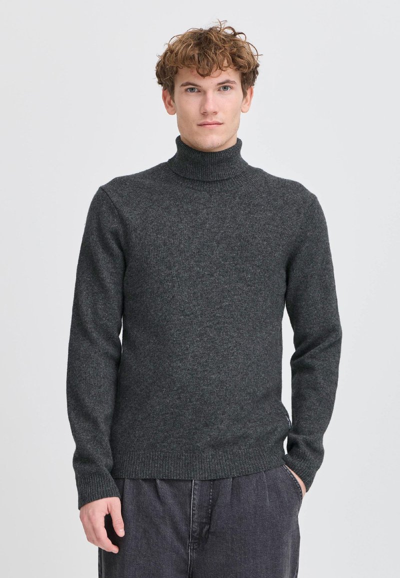 Giovane uomo con capelli ricci che indossa un maglione a collo alto grigio scuro e pantaloni scuri, in piedi davanti a uno sfondo chiaro e semplice.