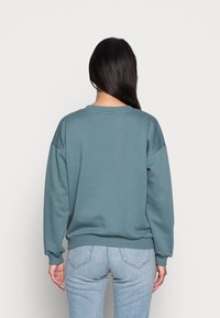 Sweat-shirt en polaire dans un ton teal atténué, avec un col rond, des épaules tombantes et des poignets et ourlet côtelés. Vue de dos montrant une texture lisse.