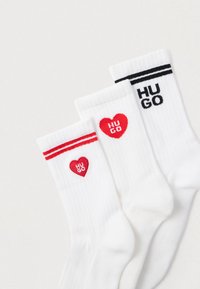 Tre par hvide sokker med forskellige designs: et par med et rødt hjerte-logo med "HUGO," et andet med røde striber, og det sidste med et sort logo.