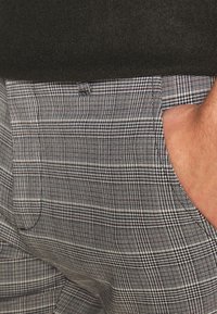 Main dans la poche d’un pantalon à carreaux gris porté avec un pull foncé.