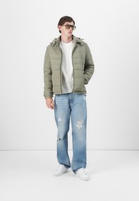 Polstret olivengrønn jakke med hette, båret over en lys grå skjorte. Lys blå slitte jeans og hvite sneakers fullfører antrekket.