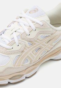 Ljust beige sportskor med mesh- och mockamaterial, vita snören och en texturerad design med en synlig stötdämpande sula.