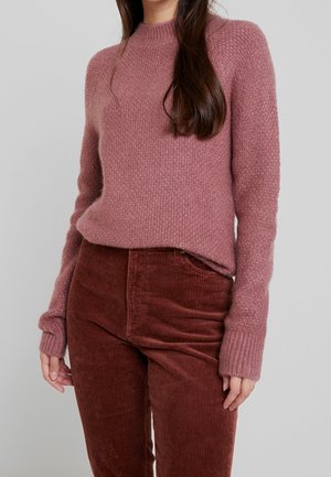 Femme portant un pull en tricot mauve avec des poignets côtelés et un pantalon en velours côtelé marron taille haute, debout devant un fond uni.